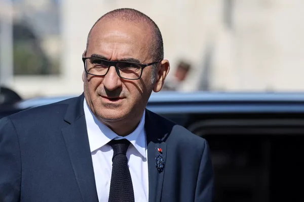 Portrait en extérieur d'un homme aux cheveux courts portant des lunettes, un costume sombre, une chemise blanche et une cravate. Il regarde légèrement de côté avec une expression neutre.
