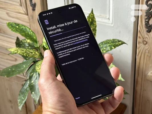 Main tenant un smartphone noir montrant l'installation d'une mise à jour de sécurité, avec une porte en bois et une plante en arrière-plan.