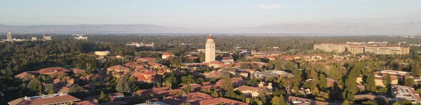 The image is an aerial view of Stanford University, featuring the campus buildings with distinctive red-tile roofs. At the center of the image is the iconic Hoover Tower, surrounded by greenery and trees. In the background, one can see the foothills and distant mountains, with a glimpse of the San Francisco Bay. The scene is set during the day under clear skies, with a wide landscape of the university's surroundings.