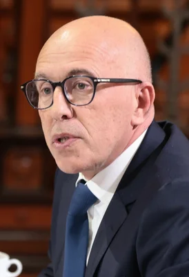 Éric Ciotti de profil, s'exprimant dans un cadre intérieur avec mobilier en bois.