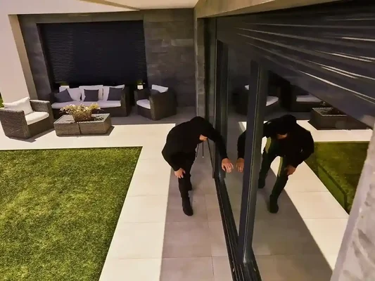 Individu cagoulé accroupi sur une terrasse devant une porte vitrée, illustrant un home-jacking.
