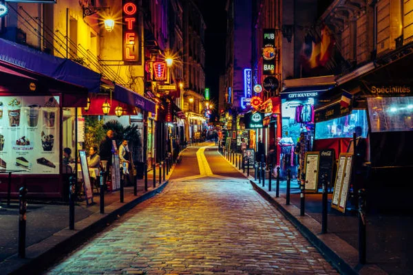 Rue nocturne typique de Paris, avec ses pavés et ses enseignes lumineuses.