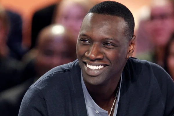 Omar Sy souriant, cardigan bleu et chaîne en or, en public.