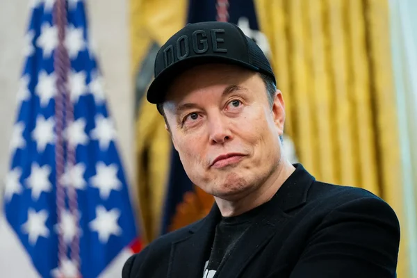 Elon Musk portant une casquette 'DOGE' devant un drapeau américain.