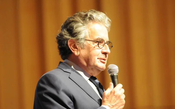Alain Claeys de profil, micro à la main, devant un rideau orange.