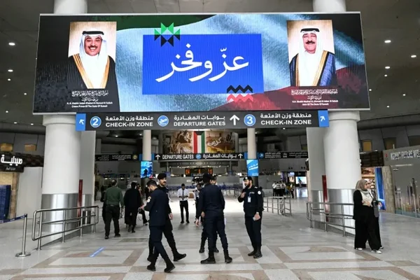 Aéroport au Koweït : un écran géant affiche les portraits des dirigeants, tandis que des voyageurs et agents de sécurité circulent.
