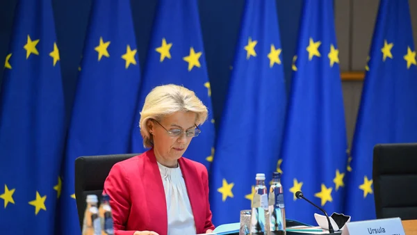 Femme en blazer rose assise à une table de conférence devant des drapeaux de l'UE.