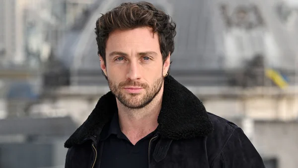 Aaron Taylor-Johnson en veste noire à col en shearling dans un décor urbain.