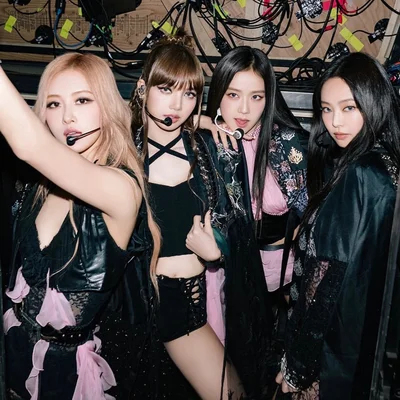 Blackpink en coulisses, en costumes de scène et micros-casques, posant devant un mur technique.