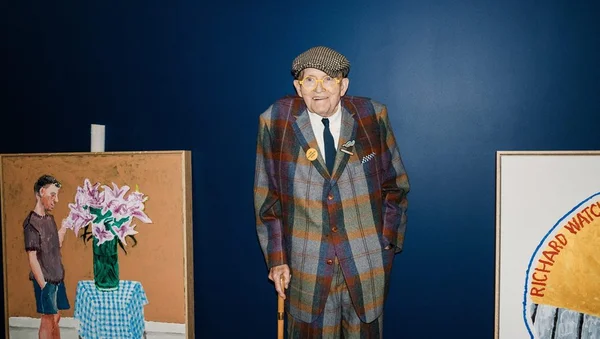 David Hockney posant avec sa canne entre deux de ses tableaux.