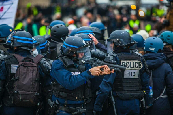 Groupe de policiers français en tenue anti-émeute, casques et gilets pare-balles, lors d'une manifestation ; un officier au premier plan vise avec un lanceur de projectiles non létaux.