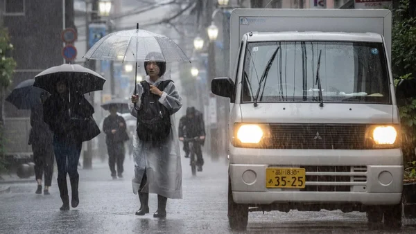 Un piéton sous une forte pluie dans une rue japonaise, avec un camion de livraison roulant sur la chaussée détrempée.