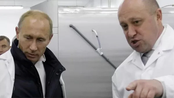 Vladimir Poutine en visite dans un laboratoire ou une installation industrielle, écoutant un interlocuteur en blouse blanche.