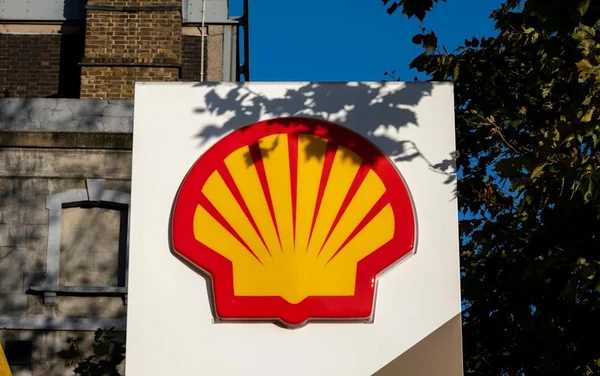 Logo de Shell sur un panneau extérieur, avec ciel bleu et feuillage.