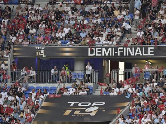 Foule enthousiaste dans les tribunes lors d'une demi-finale du Top 14, avec drapeaux et banderoles rouges.