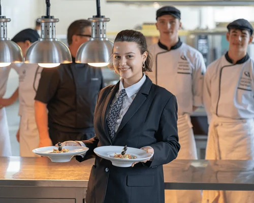 Jeune femme en costume tenant des assiettes dans une cuisine professionnelle, entourée de chefs.