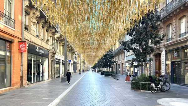 Rue commerçante toulousaine ombragée par une canopée de rubans dorés, mesure de rafraîchissement estival.
