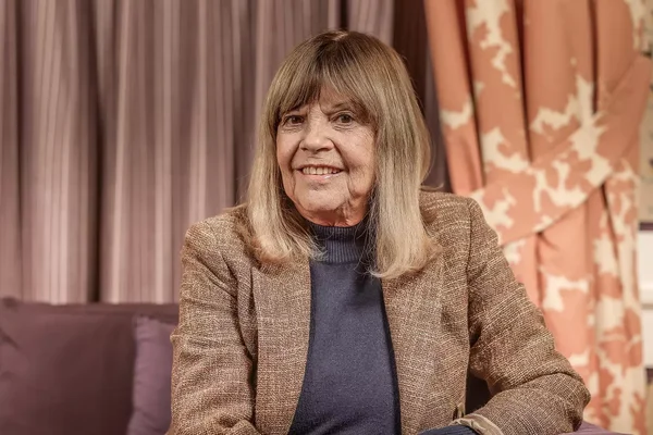 Chantal Goya, souriante, assise devant des rideaux mauves en veste en tweed et col roulé bleu.
