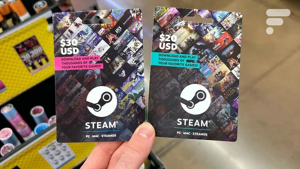 Une main tenant deux cartes Steam physiques, d'une valeur de 30 et 20 dollars, dans un magasin.