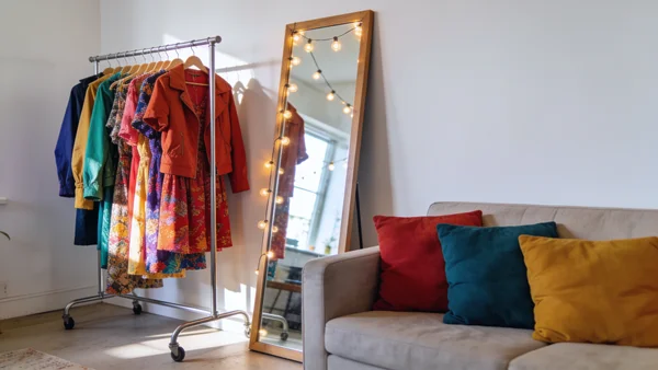 Portant métallique avec des robes et vestes suspendues, miroir posé au sol, guirlande lumineuse accrochée au mur, canapé avec coussins dans un salon cosy
