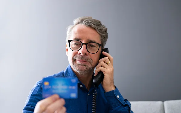 Un homme utilisant un téléphone fixe tout en tenant une carte de crédit, illustration générique d'une fraude téléphonique.