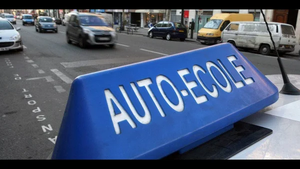 Panneau 'AUTO-ECOLE' bleu sur un véhicule, arrière-plan urbain flou.