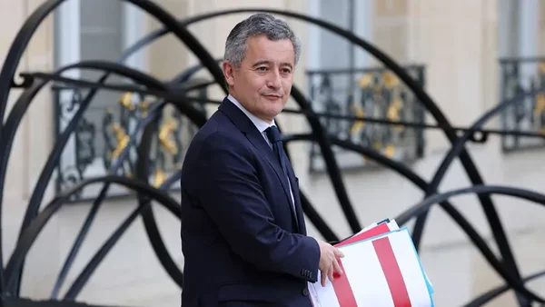 Gérald Darmanin à l'extérieur, tenant un dossier, devant un bâtiment officiel.