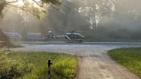 Hélicoptère stationné dans un cadre boisé, avec des véhicules à l'arrière-plan.