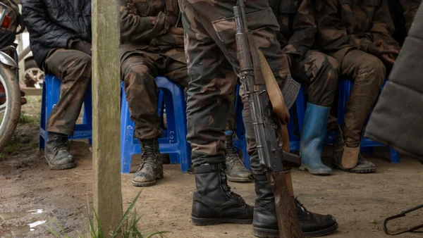 Soldats armés assis sur des tabourets bleus, un garde en faction avec un fusil AK au premier plan.