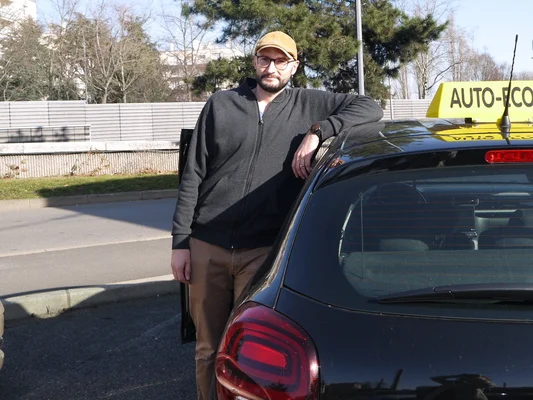 Homme en casquette jaune appuyé contre une voiture noire portant un panneau 'AUTO-ECO'.
