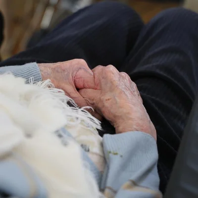 Mains jointes d'une personne âgée sur ses genoux, couvertes d'une couverture blanche.