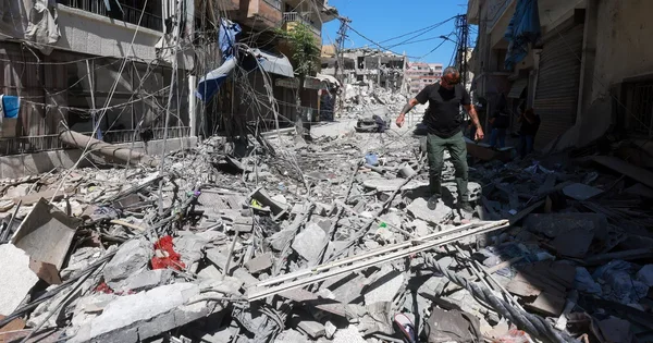 Un homme traverse une rue couverte de gravats et de débris après des destructions, sous un ciel dégagé.