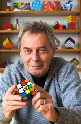 Ernő Rubik souriant, entouré de puzzles sur des étagères.