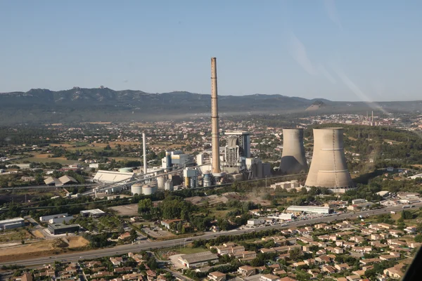 Vue aérienne de la centrale biomasse de Gardanne, avec ses tours de refroidissement et sa cheminée, près d'un quartier résidentiel et des montagnes.