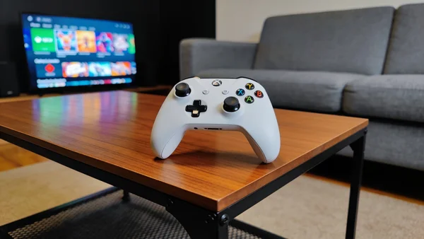 Manette Xbox blanche posée sur une table basse en bois, à côté d'un canapé gris, lumière tamisée du salon, écran de télévision allumé au loin montrant un menu de jeu