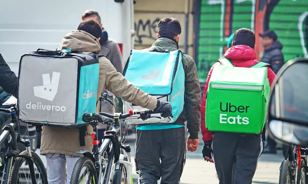Livreurs à vélo des plateformes Deliveroo et Uber Eats, vus de dos, marchant avec leurs bicyclettes.