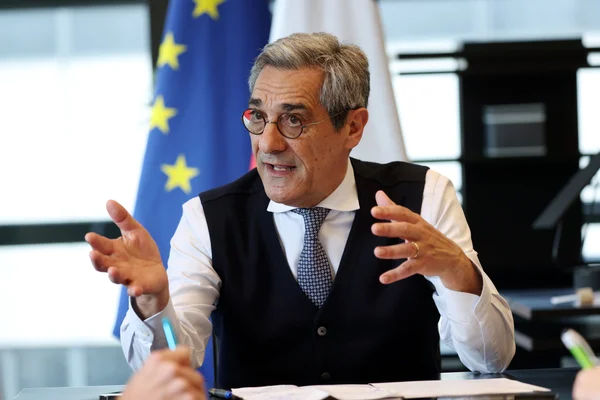 Le ministre Serge Papin en interview, gesticulant devant le drapeau européen.