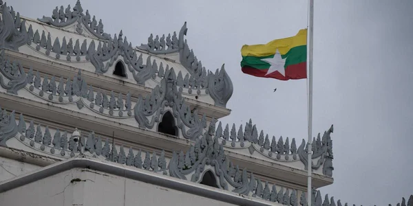 Drapeau birman flottant devant un bâtiment officiel au toit traditionnel.