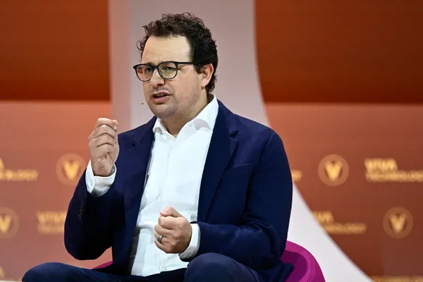 Portrait d'un intervenant s'exprimant lors d'une conférence ou d'un entretien, avec des logos en arrière-plan.
