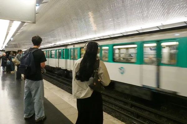 Voyageurs sur un quai de métro parisien, un train en mouvement en arrière-plan.