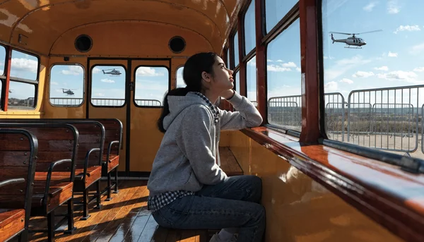 Adolescente assise seule dans un bus scolaire, regardant par la fenêtre des barrières métalliques et des hélicoptères dans le ciel, expression anxieuse, dans une atmosphère de stress et d'attente