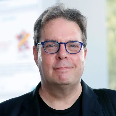 Gros plan de Douglas Kennedy, lunettes bleues, veste noire, fond flou.