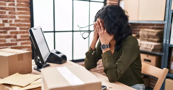 Une femme en détresse au travail, se cachant le visage entre ses mains tout en tenant ses lunettes, assise à un bureau dans un entrepôt