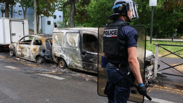 Un policier français en tenue anti-émeute monte la garde à côté d'un fourgon et d'une voiture calcinés sur une rue bordée d'arbres, témoignant de la violence des émeutes.