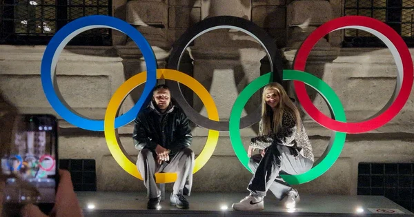 Dévoilement des emblèmes des JO Alpes françaises 2030 à Briançon : un homme et une femme posent sur les anneaux olympiques lumineux, photographiés par un spectateur.