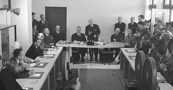 Réunion officielle lors des accords d'Évian, avec un homme debout s'adressant aux délégués et photographes.