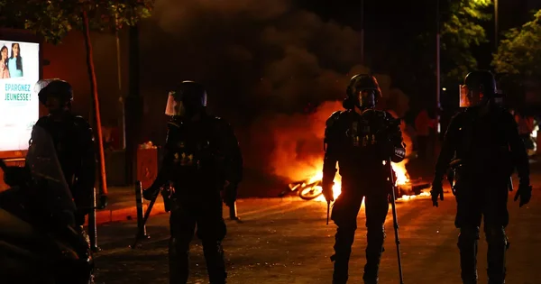 Quatre policiers en tenue anti-émeute se tiennent sur une rue la nuit, silhouettés contre un grand incendie qui brûle en arrière-plan. Un panneau publicitaire avec du texte français est visible sur la gauche.