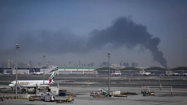 Tarmac d'aéroport avec des avions d'Emirates et une fumée noire au loin, illustrant les tensions géopolitiques dans la région.