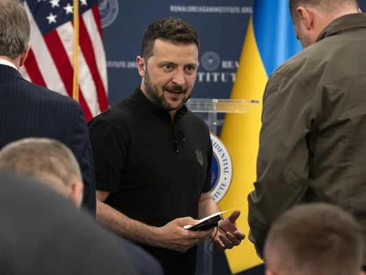 Volodymyr Zelenskyy en discussion lors d'un sommet, avec les drapeaux américain et ukrainien en arrière-plan.
