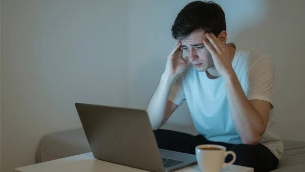 Jeune homme assis devant un ordinateur portable dans une chambre modeste, mains sur les tempes, visage fatigué, tasse de café vide, lumière de l'écran éclairant son visage